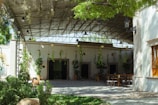 A covered outdoor space with hanging plants and vines suspended from a metal structure. The area includes wooden and metal tables and chairs, surrounded by greenery and a paved courtyard. The overhead metal roof provides shade and features several light fixtures. A wall with a single wooden-framed window and additional plants adorns the background.