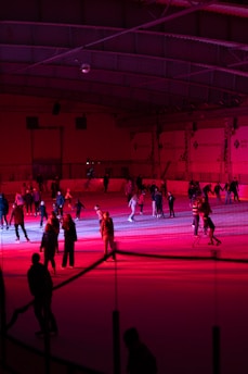 People of various ages are enjoying ice skating in an indoor rink. The environment is illuminated with vibrant red and pink lighting, creating a vivid and lively atmosphere. Skaters are scattered across the ice, some moving in groups while others appear to be learning with aids.