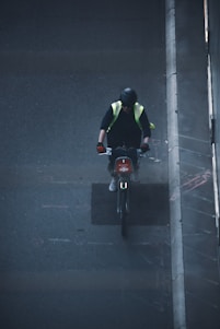 A person wearing a black helmet and a high-visibility vest is riding a bicycle on a dark, urban street. They have gloves on their hands and appear to be focused on the road ahead. The image is taken from an elevated perspective, possibly from a bridge or building, showing part of the sidewalk and the urban environment.