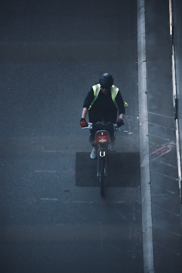 A person wearing a black helmet and a high-visibility vest is riding a bicycle on a dark, urban street. They have gloves on their hands and appear to be focused on the road ahead. The image is taken from an elevated perspective, possibly from a bridge or building, showing part of the sidewalk and the urban environment.