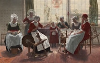 A group of women dressed in traditional clothing sit and engage in conversation in a cozy room, with a spinning wheel and a young girl in the center. The room is warmly lit, with large windows in the background allowing natural light to fill the space. Some women are seated on chairs, while others stand or lean in to participate.