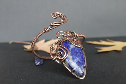 A handcrafted pendant featuring an intricate wire design that holds a large blue stone with white marbling. The wire is a copper color and is artistically wound around the stone with spirals and loops. The background includes two blurred autumn leaves set against a dark surface.