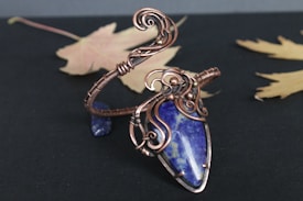 A handcrafted wire-wrapped pendant featuring a blue gemstone as the centerpiece. The intricate copper wire designs encase the stone, creating an elaborate and artistic look. The background consists of a dark surface with a few faded autumn leaves placed around the pendant.