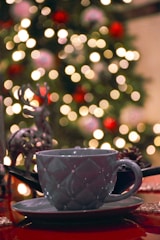 A ceramic mug with a textured design, resting on a matching saucer on a table. In the background, a Christmas tree is adorned with colorful ornaments and blurred festive lights, creating a bokeh effect. There is a decorative reindeer figure next to the mug, enhancing the holiday theme.