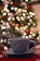 A ceramic mug with a textured design, resting on a matching saucer on a table. In the background, a Christmas tree is adorned with colorful ornaments and blurred festive lights, creating a bokeh effect. There is a decorative reindeer figure next to the mug, enhancing the holiday theme.