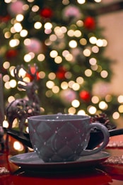 A ceramic mug with a textured design, resting on a matching saucer on a table. In the background, a Christmas tree is adorned with colorful ornaments and blurred festive lights, creating a bokeh effect. There is a decorative reindeer figure next to the mug, enhancing the holiday theme.