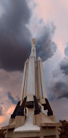 A large rocket stands vertically, reaching towards a dramatic sky filled with dark, swirling clouds. The base of the rocket is supported by a solid structure, and the evening light casts shadows, enhancing its imposing presence.