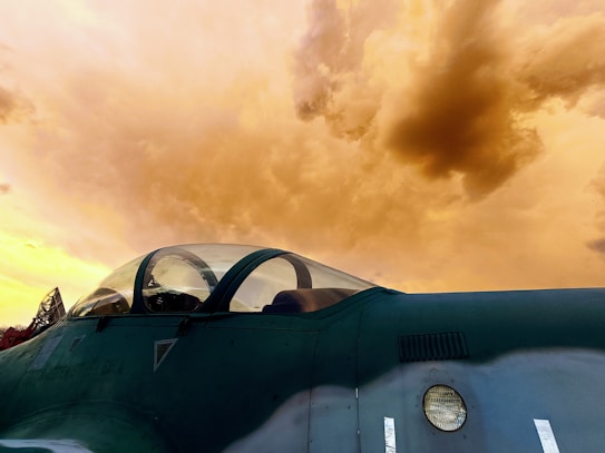 A close-up view of an aircraft cockpit against a dramatic, cloudy sky. The cockpit features a canopy with a reflective surface, and the aircraft's body is painted in a dark green color. The clouds in the sky are tinted orange, suggesting a sunset or sunrise setting.