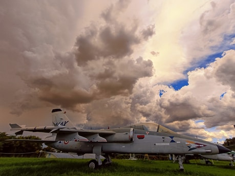 A grounded military jet with various markings and symbols is stationed on a grassy area under a dramatic sky filled with large, dark clouds. The lighting suggests an impending storm, with a mixture of golden tones and patches of bright blue sky peeking through.