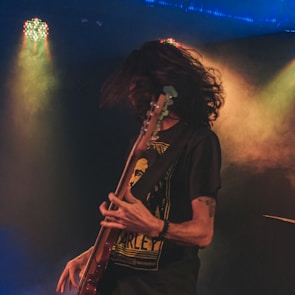 A musician with long hair is passionately playing an electric guitar on stage, with dynamic lighting and smoke creating a dramatic atmosphere. The person is wearing a dark t-shirt with a graphic design.