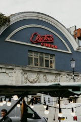 A classic cinema facade features an arched roof with a neon sign spelling 'Electric Cinema' in cursive red letters. The building's exterior is adorned with intricate floral carvings and has a vintage architectural style. In the foreground, out-of-focus decorative bulbs hang, adding a cozy ambiance, while a street vendor's stand and some parts of vehicles are partially visible.