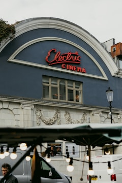 A classic cinema facade features an arched roof with a neon sign spelling 'Electric Cinema' in cursive red letters. The building's exterior is adorned with intricate floral carvings and has a vintage architectural style. In the foreground, out-of-focus decorative bulbs hang, adding a cozy ambiance, while a street vendor's stand and some parts of vehicles are partially visible.