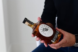A bottle of whiskey is held in someone's hands. The label on the bottle is clearly visible, displaying graphics and text. The person is wearing a dark long-sleeved shirt and a watch on their wrist.