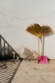 A broom with a yellow handle is resting against a white wall beside a pink dustpan. The wall has visible cracks and mold at the bottom, creating a weathered look. A shadow from a nearby railing falls across the concrete floor, adding dimension to the scene.