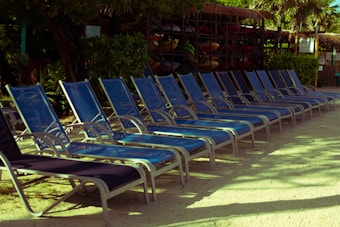A row of empty blue lounge chairs is aligned on a sandy surface, partially shaded by surrounding trees and vegetation. In the background, there are wooden structures and storage racks filled with colorful kayaks, adding a sense of leisure and outdoor activity.