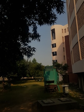 A green mechanical structure is situated on a grassy area near a modern, multi-story building with round edges and perforated architectural details. The scene is shadowed by tree foliage in the foreground, and there are more trees in the background.