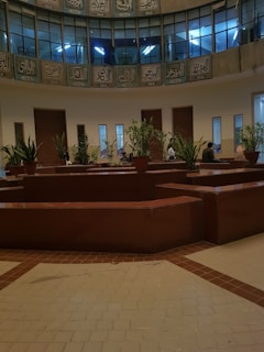 A spacious indoor area features a circle of potted plants placed on brown, geometrically shaped counters. Above, Arabic script adorns the circular balcony with a series of windows showing blue-lit interiors. Several small groups of people can be seen seated around the plants, adding vitality to the serene setting.