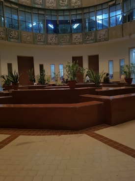 A spacious indoor area features a circle of potted plants placed on brown, geometrically shaped counters. Above, Arabic script adorns the circular balcony with a series of windows showing blue-lit interiors. Several small groups of people can be seen seated around the plants, adding vitality to the serene setting.