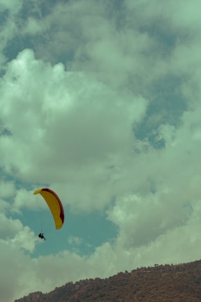 A person is paragliding with a yellow and red parachute high in the sky against a backdrop of fluffy clouds. Below, a mountain range is visible, indicating an outdoor adventure setting.