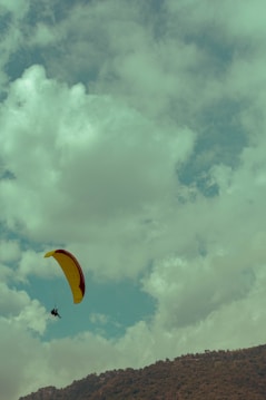 A person is paragliding with a yellow and red parachute high in the sky against a backdrop of fluffy clouds. Below, a mountain range is visible, indicating an outdoor adventure setting.