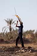 A person stands outdoors aiming a bow and arrow upwards. The individual is wearing a dark outfit and appears to be in the middle of a rural area with sparse vegetation and palm trees in the background. The ground is dry with some rocks scattered around.