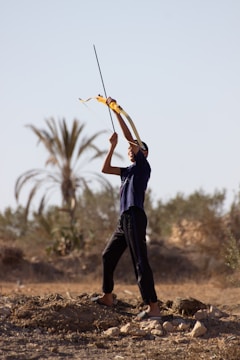 A person stands outdoors aiming a bow and arrow upwards. The individual is wearing a dark outfit and appears to be in the middle of a rural area with sparse vegetation and palm trees in the background. The ground is dry with some rocks scattered around.