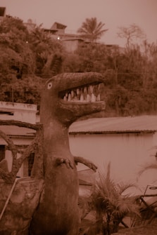 A large dinosaur sculpture with an open mouth and visible teeth stands in an outdoor setting, surrounded by foliage and buildings. The sepia tone gives the scene a vintage or dated appearance.