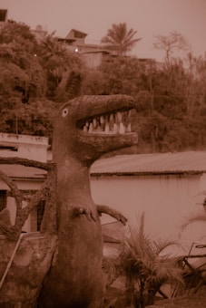 A large dinosaur sculpture with an open mouth and visible teeth stands in an outdoor setting, surrounded by foliage and buildings. The sepia tone gives the scene a vintage or dated appearance.