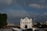 A classic white colonial-style church is positioned prominently in the foreground, surrounded by a mix of modern and traditional buildings. A faint rainbow arcs across the cloudy sky above, adding a serene and hopeful atmosphere to the scene. Communication towers are visible, and lush greenery is evident on the left.