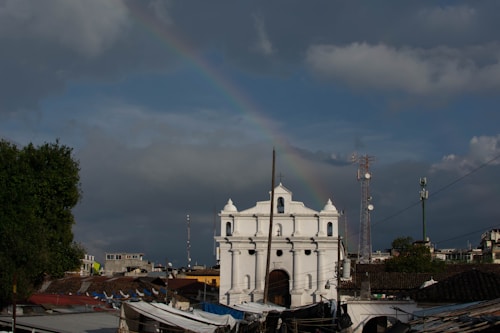 A classic white colonial-style church is positioned prominently in the foreground, surrounded by a mix of modern and traditional buildings. A faint rainbow arcs across the cloudy sky above, adding a serene and hopeful atmosphere to the scene. Communication towers are visible, and lush greenery is evident on the left.