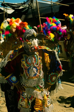 A person dressed in a vibrant and intricately detailed traditional costume, featuring colorful beadwork, tassels, and ornate designs. The headpiece is adorned with a mask and decorative elements. In the background, other similarly dressed participants are visible in a festive setting.