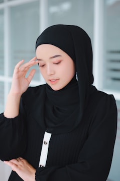 A person wearing a black hijab and a black outfit with white buttons stands with one hand gently touching their temple, eyes closed, conveying a serene expression against a neutral background.