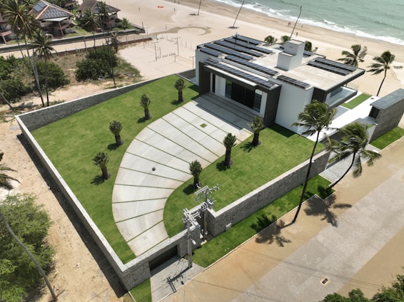 A modern house with a flat roof and solar panels is situated near a beach. The property is surrounded by a stone wall and features a well-manicured lawn with several small palm trees. There is a paved driveway leading to the entrance, and the beach is visible with gentle waves meeting the sand. Palm trees are scattered along the property and the beach.