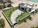 A modern house with a flat roof and solar panels is situated near a beach. The property is surrounded by a stone wall and features a well-manicured lawn with several small palm trees. There is a paved driveway leading to the entrance, and the beach is visible with gentle waves meeting the sand. Palm trees are scattered along the property and the beach.