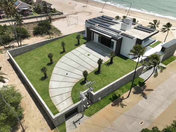 A modern house with a flat roof and solar panels is situated near a beach. The property is surrounded by a stone wall and features a well-manicured lawn with several small palm trees. There is a paved driveway leading to the entrance, and the beach is visible with gentle waves meeting the sand. Palm trees are scattered along the property and the beach.