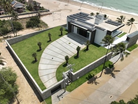 A modern house with a flat roof and solar panels is situated near a beach. The property is surrounded by a stone wall and features a well-manicured lawn with several small palm trees. There is a paved driveway leading to the entrance, and the beach is visible with gentle waves meeting the sand. Palm trees are scattered along the property and the beach.