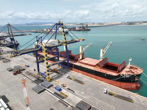A large cargo ship is docked at a port alongside massive cranes used for loading and unloading. The ship is painted black with red accents and has open cargo holds visible on its deck. Several trucks are positioned along the dock, possibly for transporting goods. The surrounding water is a bright turquoise, and the horizon shows distant landforms under a partly cloudy sky.