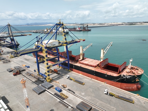 A large cargo ship is docked at a port alongside massive cranes used for loading and unloading. The ship is painted black with red accents and has open cargo holds visible on its deck. Several trucks are positioned along the dock, possibly for transporting goods. The surrounding water is a bright turquoise, and the horizon shows distant landforms under a partly cloudy sky.