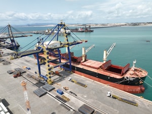 A large cargo ship is docked at a port alongside massive cranes used for loading and unloading. The ship is painted black with red accents and has open cargo holds visible on its deck. Several trucks are positioned along the dock, possibly for transporting goods. The surrounding water is a bright turquoise, and the horizon shows distant landforms under a partly cloudy sky.