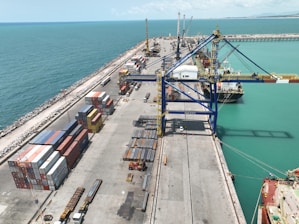 A bustling port with stacks of shipping containers neatly arranged on the dock. Large cranes stand ready for loading and unloading vessels. The seawater appears turquoise, and a few ships are docked, with workers and vehicles visible on the ground.