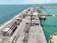 A bustling port with stacks of shipping containers neatly arranged on the dock. Large cranes stand ready for loading and unloading vessels. The seawater appears turquoise, and a few ships are docked, with workers and vehicles visible on the ground.