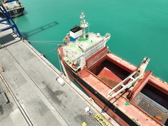 A large cargo ship is docked at a port with an open cargo hold, positioned parallel to a concrete pier. The turquoise water contrasts with the red and white colors of the ship. A crane or loading device is visible onboard the ship, indicating loading or unloading activity. Various vehicles and equipment are seen on the dock.