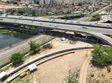 An elevated highway with multiple lanes is busy with traffic, including cars and motorcycles. Below the highway, a construction site features long pipes and workers. A river runs next to railway tracks that parallel the road. In the background, there are residential buildings and greenery.