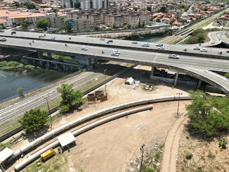 An elevated highway with multiple lanes is busy with traffic, including cars and motorcycles. Below the highway, a construction site features long pipes and workers. A river runs next to railway tracks that parallel the road. In the background, there are residential buildings and greenery.