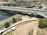 An elevated highway with multiple lanes is busy with traffic, including cars and motorcycles. Below the highway, a construction site features long pipes and workers. A river runs next to railway tracks that parallel the road. In the background, there are residential buildings and greenery.