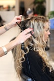 A hair stylist is curling the long blonde hair of a seated woman using a curling iron. The woman appears to be in a salon setting, wearing a black styling cape. The stylist's hands are in focus, showing red nail polish and holding the curling iron.