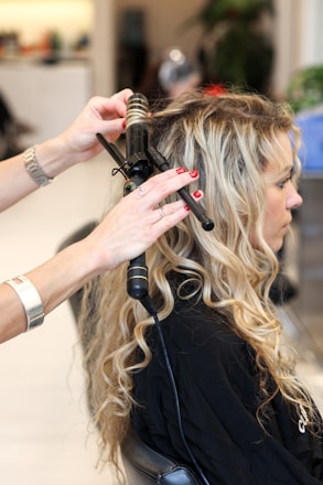 A hair stylist is curling the long blonde hair of a seated woman using a curling iron. The woman appears to be in a salon setting, wearing a black styling cape. The stylist's hands are in focus, showing red nail polish and holding the curling iron.