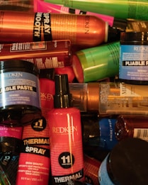 A collection of various hair styling products from the brand Redken, including thermal sprays, pliable paste, and other haircare items in a variety of colorful packaging such as red, orange, blue, and green bottles and jars.