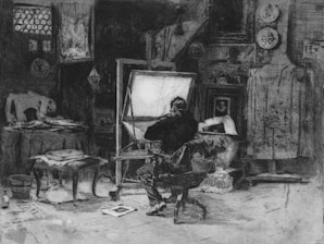 An artist's studio with a man seated in front of an easel, surrounded by various artworks and clutter. The room is filled with framed paintings and drawings hung on the walls, a table covered in papers and a coat is draped over a chair. The setting is dimly lit, creating a sense of depth and atmosphere.