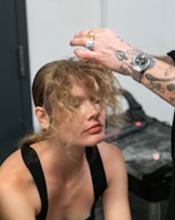 A woman with curly hair is receiving a hair touch-up from a person with tattooed arms and rings. The setting appears to be indoors, possibly a salon or dressing room, with a gray countertop visible in the background.